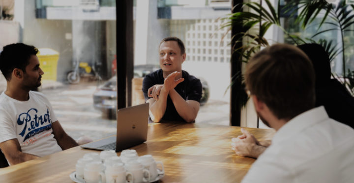 In einem hellen Raum der KI Agentur trifft sich eine Gruppe von Menschen um einen Holztisch. Eine Person gestikuliert beim Sprechen, die anderen hören aufmerksam zu. Auf dem Tisch steht ein geöffneter Laptop, daneben mehrere Tassen. Im Hintergrund sind große Fenster zu sehen.